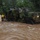 A military truck in camouflage paint is navigating through deep, fast-moving floodwaters on a flooded road or street. A soldier in uniform is visible leaning out of the cab area of the vehicle. The scene appears to be a flood rescue or emergency response operation, with additional personnel visible in the background near a building.