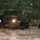 A military heavy tactical truck (appears to be an M977 HEMTT or similar) is navigating through deep flood waters on a submerged road or street. Rescue personnel in yellow dry suits and helmets are wading through the muddy brown floodwater alongside the vehicle. The scene depicts an active flood rescue or evacuation operation in a residential or wooded area.