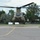 A military CH-47 Chinook tandem-rotor helicopter is hovering just above a concrete helipad, appearing to be in the process of landing or taking off. The helicopter is painted in olive drab/army green camouflage and is viewed from a front-facing perspective. The setting appears to be a civilian or semi-military area with trees, a fence line, and bystanders visible in the background.