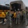 Military personnel in US Army camouflage uniforms are conducting what appears to be an emergency evacuation operation alongside a Boulder Valley School District school bus. Civilians, including at least one person with a walker and individuals with dogs, are being assisted from a military transport truck. The scene takes place in rainy conditions, suggesting a disaster relief or flood evacuation scenario.