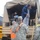 Military personnel in ACU (Army Combat Uniform) camouflage are assisting civilians disembarking from a canvas-covered military transport truck. One civilian is wearing pajamas/sleepwear, suggesting an emergency evacuation scenario. The scene appears to take place in cold, snowy conditions with safety personnel wearing high-visibility orange vests also present.