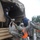 Military personnel in US Army ACU (Army Combat Uniform) camouflage are assisting civilians onto or off of a military tactical truck during what appears to be a disaster relief or evacuation operation. The scene takes place in rainy conditions, with soldiers wearing high-visibility orange safety vests helping people climb a wooden ladder into the canvas-covered truck bed. A civilian in a dark cap is visible inside the truck receiving a bag being passed up.