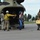 Civilians are being evacuated via a large military helicopter, likely a CH-47 Chinook, on a tarmac. A smiling woman with a backpack exits the aircraft while a person in a yellow firefighting jacket assists near the ramp. Another individual carries a yellow pet carrier, suggesting this may be a wildfire or disaster evacuation operation.