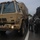 A military truck marked 'COARNG 193MP' and 'HC202' is parked on a wet road alongside a yellow school bus numbered 231, suggesting a disaster relief or evacuation operation during rainy conditions. Personnel in rain gear are visible between the vehicles, apparently coordinating or transferring people or supplies. The scene appears to involve National Guard forces assisting with a civilian emergency, likely a hurricane or flood event.