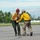 Two wildland firefighters in standard yellow Nomex shirts and hard hats are handling a golden/orange-colored dog on a paved surface, likely a tarmac or airstrip. One firefighter wearing a red helmet is carrying the dog while the other in a gray helmet holds a purple leash. The scene appears to be an animal rescue or evacuation operation associated with a wildfire response.