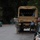 A military utility truck (LMTV or similar canvas-topped Army vehicle) is stopped on a wet residential street. Two soldiers in ACU (Army Combat Uniform) camouflage are standing near the rear of the vehicle, while a civilian in blue shirt and brown pants bends down near the rear axle area. The scene appears to involve a vehicle check or minor mechanical issue during what may be a disaster relief or civil support operation.