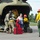 A military/emergency helicopter evacuation is underway on a tarmac, with personnel assisting a civilian woman and her dog to disembark. Multiple responders wearing yellow firefighter shirts, green pants, and red helmets are present, suggesting this is likely a wildfire evacuation operation. Luggage including a red suitcase and a dark travel bag are visible on the ground near the helicopter's open rear cargo door.