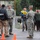 The image shows a multi-agency emergency response scene on a wet street, likely during or after a flood or natural disaster. Military personnel in ACU camouflage uniforms are coordinating with a Sheriff's deputy and civilians, including a person carrying a young child in a yellow rain jacket. Orange traffic cones are placed on the road, and military vehicles are visible in the background.