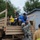 A military evacuation operation is underway, with civilians including children and a baby being helped off a large tan military truck. Personnel from the Lyons Fire Protection District (Lyons FPD) and U.S. Army soldiers in camouflage uniforms are assisting evacuees. This appears to be a disaster relief or emergency evacuation scenario, likely related to flooding given the context of Lyons, Colorado.