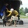 A rescue or evacuation operation is underway at what appears to be a small airfield or helipad. A firefighter or emergency responder in a yellow shirt and white hard hat is unloading a pet carrier and managing a black dog on a leash from a military-style helicopter. A soldier in camouflage uniform and a civilian in a blue jacket are also present near the aircraft.