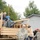 Military personnel and civilians are engaged in what appears to be an evacuation or rescue operation using a tan military cargo truck. Soldiers in ACU (Army Combat Uniform) camouflage are assisting civilians boarding the flatbed vehicle, which already has people and luggage loaded on it. The wet pavement and overcast sky suggest recent or ongoing adverse weather conditions, consistent with a flood or disaster response scenario.