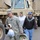 A U.S. Army soldier in ACU (Army Combat Uniform) is assisting civilians, carrying a baby in an infant car seat along with a plastic bag of supplies. Two civilians, a woman wearing a 'Wyoming' sweatshirt and a man in a dark hoodie and blue cap, follow behind. A large tan military tactical vehicle (LMTV or similar) is visible in the background, suggesting a disaster relief or evacuation operation.