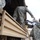 Military personnel in US Army ACU (Army Combat Uniform) digital camouflage are loading or unloading equipment onto/from a tan military flatbed truck. One soldier is positioned on the truck bed while another stands on the ground handling what appears to be a gray plastic equipment case or container. A wooden-slatted ladder is propped against the truck to assist with access.
