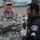 Two uniformed personnel are engaged in a friendly conversation outdoors. One individual is wearing U.S. Army ACU (Army Combat Uniform) with a military cap, while the other is wearing a Boulder County Sheriff's Office jacket with sergeant chevrons. Both appear relaxed and smiling, suggesting a cooperative inter-agency interaction.