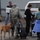 A group of four people and a dog are gathered in a parking area near what appears to be a Boulder Sheriff's vehicle. One person in military camouflage uniform is holding a dog on a leash while others handle boxes and a hand truck, suggesting a supply distribution or relief effort. The scene appears to take place in an overcast outdoor setting near a gray building.