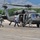 A military UH-60 Black Hawk helicopter is parked on a tarmac at what appears to be a small regional airport. Several individuals, including two soldiers in Army Combat Uniforms (ACU) and two civilians, are moving around the aircraft, suggesting a recent landing or evacuation operation. The mountainous, tree-lined background and general setting suggest a western U.S. location.