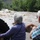 Two elderly individuals, viewed from behind, are observing a severely flooded and turbulent river from what appears to be a bridge or elevated vantage point. The river is running extremely high with brown, sediment-laden water and visible debris including uprooted trees. The surrounding landscape shows dense green vegetation and hills in the background.
