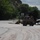 A military-style truck carries multiple civilians through a flood-damaged road covered in mud and debris. The scene appears to be a post-flood evacuation or rescue operation in a small Colorado town, with visible road damage, displaced debris, and a damaged vehicle nearby. Rocky terrain and dense trees are visible in the background.