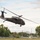 A military helicopter, consistent with a UH-60 Black Hawk, is captured in low-altitude flight over what appears to be a small airfield or open ground area. The aircraft is painted in a dark olive/black military color scheme and is flying at a low elevation above the terrain. The background features trees, utility poles, power lines, and distant hills under a partly cloudy sky.