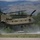 A United States Army CH-47 Chinook tandem-rotor helicopter is captured in a low hover or takeoff/landing phase at what appears to be a small airfield or helipad. The rotor wash is visibly kicking up dust and debris from the ground surface below. A mountainous, tree-lined landscape forms the background, suggesting a western U.S. location.