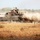 Exercise Gold Eagle smooth ride for Aussie, Marine tanks An M1 Abrams main battle tank marked '22P' is moving at speed across a dry, arid grassland terrain, kicking up a large dust cloud from its rear. A single crew member is visible in the commander's hatch position atop the turret. The environment appears to be an Australian outback training area, consistent with joint military exercises.