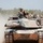 Exercise Gold Eagle smooth ride for Aussie, Marine tanks A main battle tank, identifiable as an M1 Abrams, is moving along a dusty dirt road in an arid, scrubland environment. Two crew members are visible in open hatches on top of the turret, wearing helmets and protective eyewear. The tank appears to be on a training exercise or road movement, kicking up a dust cloud behind it.