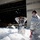 Flurry of foam released at Travis Two individuals in military/firefighting protective suits are standing in a large accumulation of fire suppression foam outside what appears to be an aircraft hangar. One person is wearing a respirator/gas mask and has their hands raised to their head, while the other is wearing sunglasses and appears to be observing. Foam particles are visibly airborne throughout the scene, suggesting an active or recently discharged foam suppression system.