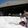 Flurry of foam released at Travis A worker in protective gear operates a red machine to clear large amounts of white foam outside a large industrial hangar marked '818'. The foam appears to be fire suppression or firefighting foam (AFFF), likely from a training exercise or accidental discharge. The scene takes place on a clear, sunny day in what appears to be a military or aviation facility.