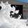 Flurry of foam released at Travis A worker in a high-visibility vest operates equipment amid a massive discharge of white firefighting foam outside a large industrial hangar numbered '818'. The foam has accumulated heavily on the ground and is actively billowing into the air, suggesting a foam system test or accidental discharge. The scene appears to take place at a military or aviation facility based on the hangar's design.