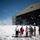 Flurry of foam released at Travis A group of approximately 8-10 individuals, including military personnel in camouflage uniforms and civilians, are standing in front of a large aircraft hangar numbered '818' that is discharging massive amounts of fire suppression foam. The foam is billowing outward and upward, covering the ground and filling the air around the group. This appears to be either an accidental foam system discharge or a controlled test of the hangar's fire suppression system.