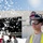 Flurry of foam released at Travis A U.S. Air Force firefighter identified as A. Stoddard (E46) stands in the foreground wearing a firefighter helmet and protective goggles with a high-visibility vest. Behind him, firefighting foam is being discharged in large quantities near what appears to be an aircraft hangar, with several other personnel visible in the background. This appears to be a firefighting training exercise or demonstration at a military installation.