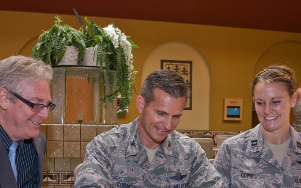 Three individuals are gathered at a counter in what appears to be a military facility. Two U.S. Air Force personnel in ABU (Airman Battle Uniform) camouflage — identified by name tapes as 'CORNISH' and 'COWIE' — are interacting with a civilian man in a suit. The central figure (CORNISH) is signing a document near a brochure titled 'Serving Our Country, Supporting Our Community.'