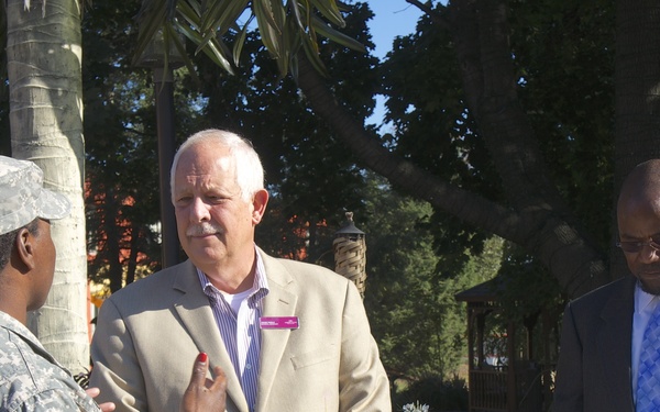 Three individuals are gathered outdoors in what appears to be a formal or ceremonial setting. A U.S. Army soldier in ACU (Army Combat Uniform) is engaged in conversation with a civilian man in a tan blazer, while a third man in a dark suit holds a folder or plaque and appears to be reading from it. The setting includes trees, landscaping, and what appears to be a building in the background.