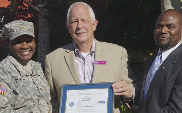 Three individuals are posing outdoors while holding a framed certificate. A U.S. Army soldier in ACU (Army Combat Uniform) stands on the left, flanked by two civilians in business attire. The certificate appears to be an ESGR (Employer Support of the Guard and Reserve) 'Patriotic Employer' award presented to 'Craig Poole' of 'Crowne Plaza'.