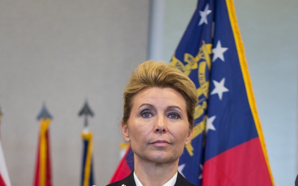 A U.S. Army officer named KUHN stands at attention in Army Service Uniform (ASU) dress blues. She wears the 82nd Airborne Division patch, aviator wings, multiple service ribbons, and rank insignia consistent with a Lieutenant Colonel. The setting appears to be a formal military ceremony with multiple flags visible in the background, including the Georgia state flag.