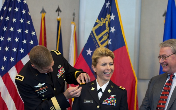 A U.S. Army promotion or award ceremony is taking place indoors. A male officer identified by nameplate as 'JARRARD' is pinning a rank insignia or device onto the shoulder of a female officer identified as 'KUNN', who is smiling broadly. A civilian man in a suit and tie stands to the right, also smiling.