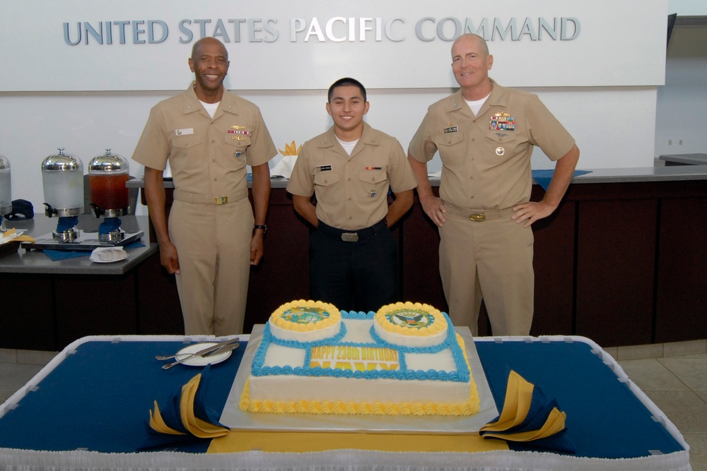 Three U.S. Navy personnel in tan/khaki uniforms are posing in front of a decorated birthday cake at the United States Pacific Command headquarters. The cake reads 'Happy 233rd Birthday,' suggesting this is a Navy birthday celebration. The individuals display varying ranks indicated by their ribbons and insignia.