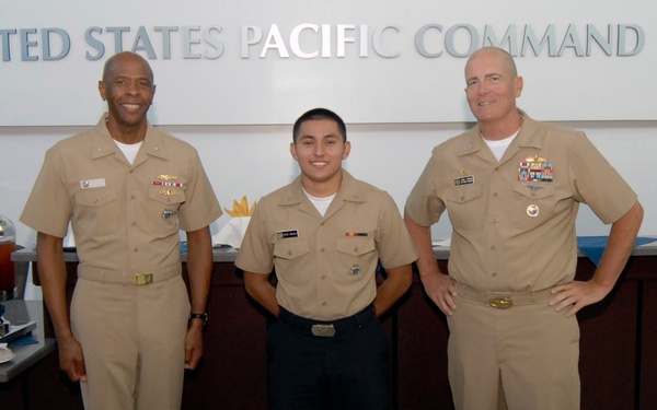Three U.S. Navy personnel in tan/khaki uniforms are posing in front of a decorated birthday cake at the United States Pacific Command headquarters. The cake reads 'Happy 233rd Birthday,' suggesting this is a Navy birthday celebration. The individuals display varying ranks indicated by their ribbons and insignia.