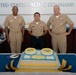 Three U.S. Navy personnel in tan/khaki uniforms are posing in front of a decorated birthday cake at the United States Pacific Command headquarters. The cake reads 'Happy 233rd Birthday,' suggesting this is a Navy birthday celebration. The individuals display varying ranks indicated by their ribbons and insignia.