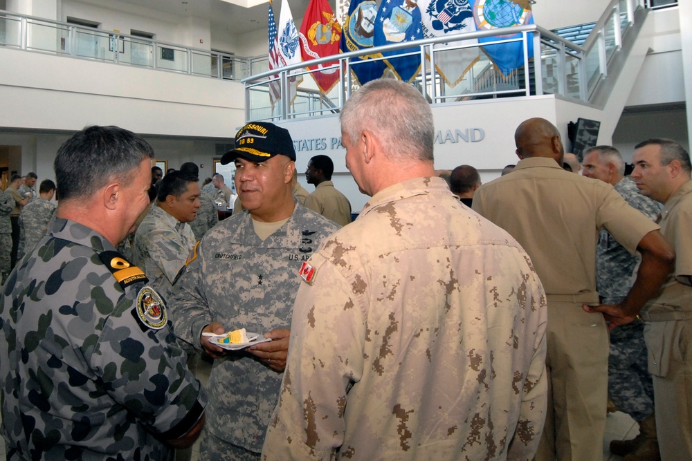 A gathering of multinational military personnel in what appears to be the United States Pacific Command headquarters, identifiable by the partial signage visible in the background. Several officers in different national uniforms are engaged in conversation, with flags of various U.S. military branches displayed on an upper level. The event appears to be a reception or informal meeting, as one officer is holding a plate with food.