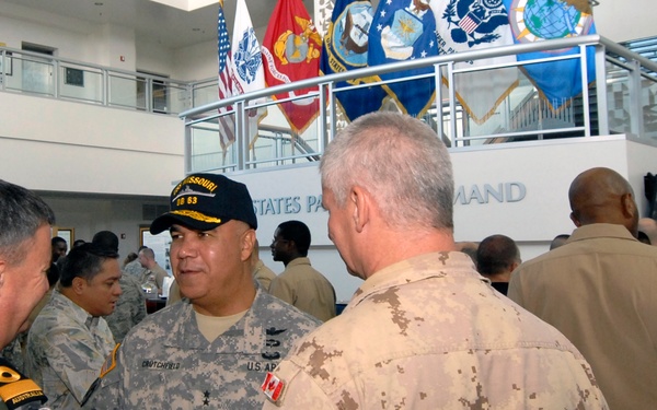 A gathering of multinational military personnel in what appears to be the United States Pacific Command headquarters, identifiable by the partial signage visible in the background. Several officers in different national uniforms are engaged in conversation, with flags of various U.S. military branches displayed on an upper level. The event appears to be a reception or informal meeting, as one officer is holding a plate with food.