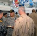 A gathering of multinational military personnel in what appears to be the United States Pacific Command headquarters, identifiable by the partial signage visible in the background. Several officers in different national uniforms are engaged in conversation, with flags of various U.S. military branches displayed on an upper level. The event appears to be a reception or informal meeting, as one officer is holding a plate with food.