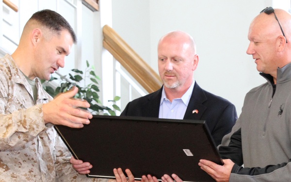 Three men are gathered indoors examining a large framed item, likely a plaque or certificate. One man is wearing a U.S. Marine Corps desert camouflage uniform (MARPAT), while the other two are in civilian attire. The setting appears to be a military administrative building or reception area.