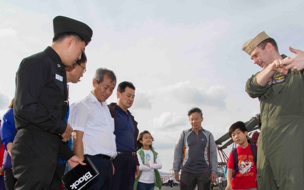 A U.S. Navy officer in a green flight suit is conducting a tour or briefing on what appears to be the flight deck of a naval vessel, gesturing toward equipment on the deck. A group of civilian visitors, including adults and children, along with a uniformed Asian military or security officer, are observing the demonstration. Helicopters are visible in the background, suggesting this is likely a public open-ship event or port visit.