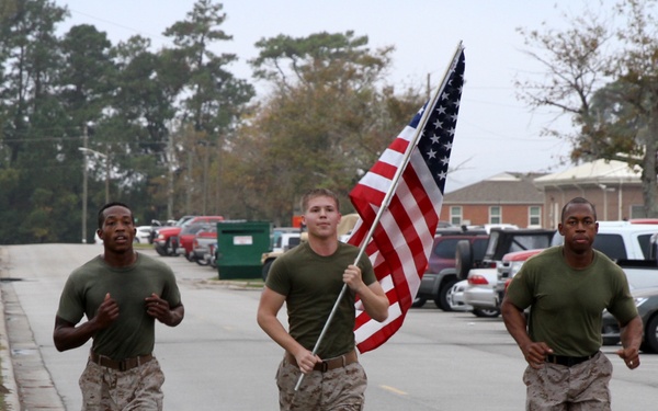 Three military personnel in U.S. Marine Corps uniforms are running along a road on what appears to be a military base. The center runner is carrying an American flag on a pole. The setting includes a parking lot with civilian vehicles and tree-lined grounds in the background.