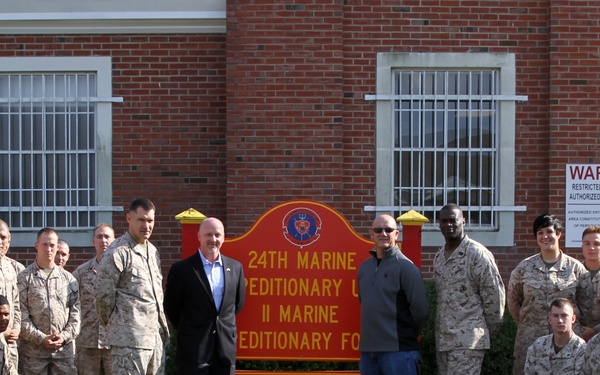 A large group of U.S. Marines in camouflage uniforms poses for a formal group photograph in front of the 24th Marine Expeditionary Unit (II Marine Expeditionary Force) headquarters building. Two civilians in dark business attire stand at the center of the group near the unit's sign. The photo appears to be an official unit or visit commemorative photograph taken outdoors on a clear day.