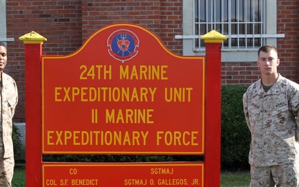 A group of six individuals, including one civilian in a suit and five U.S. Marines in uniform, pose in front of the official sign for the 24th Marine Expeditionary Unit, II Marine Expeditionary Force. The sign identifies the Commanding Officer as Col. S.F. Benedict and the Sergeant Major as SgtMaj O. Gallegos, Jr. The photo appears to be taken at a military installation, likely Camp Lejeune, North Carolina.