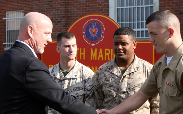 A civilian in a dark suit is shaking hands with a U.S. Marine in dress uniform, while two other Marines in desert camouflage (MARPAT) uniforms stand nearby observing. The setting appears to be outside a Marine Corps building, with a red unit sign partially visible in the background reading 'TH MARI[NE]'. This appears to be a formal greeting or recognition ceremony.