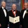 Three men in formal black tuxedos stand posed at what appears to be an AUSA (Association of the United States Army) awards ceremony. The central figure is wearing a red medallion ribbon and holding an open citation document that reads 'George Catlett Marshall Medal' awarded to 'Robert Gates'. The backdrop displays the AUSA.org logo repeatedly, and an American flag is visible on the left.