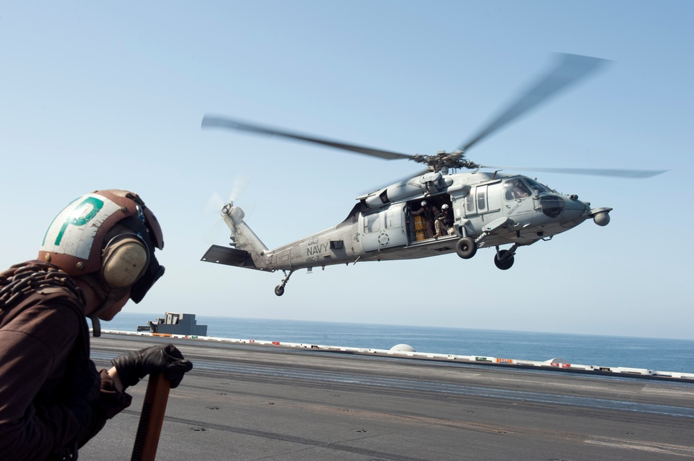 A U.S. Navy MH-60 Seahawk helicopter is hovering or taking off from the flight deck of an aircraft carrier. A flight deck crew member wearing a brown jersey and helmet with a green 'P' marking observes the helicopter from the edge of the deck. The scene appears to take place over open ocean under clear skies.